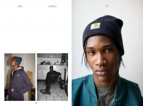 Close-up fashion editorial shot of a Black male model in a beanie, green jacket, and grey shirt against a light-grey studio backdrop.
