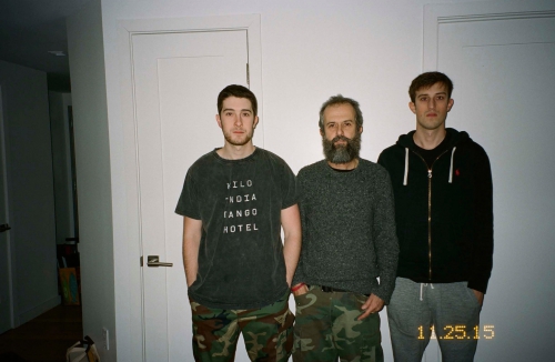 Indoor medium shot of three men of different ages standing against a white wall, wearing casual clothing, and looking at the camera.