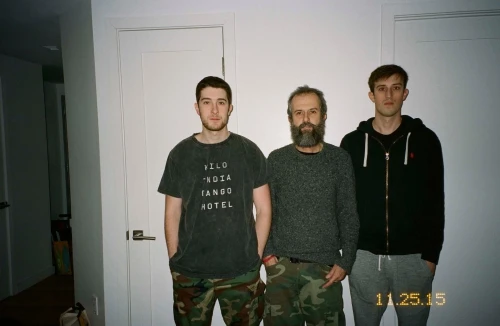 Indoor medium shot of three men of different ages standing against a white wall, wearing casual clothing, and looking at the camera.