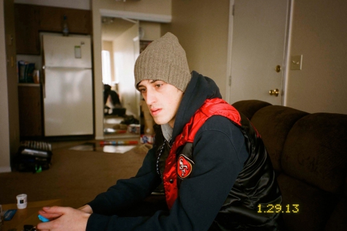 Young man wearing a grey beanie, hoodie, and red-and-black puffer vest indoors on a sofa.