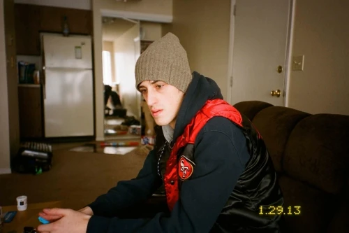 Young man wearing a grey beanie, hoodie, and red-and-black puffer vest indoors on a sofa.