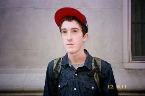 Portrait of a young adult man wearing a cap and backpack, standing in front of a building with a window. Captured in natural light.