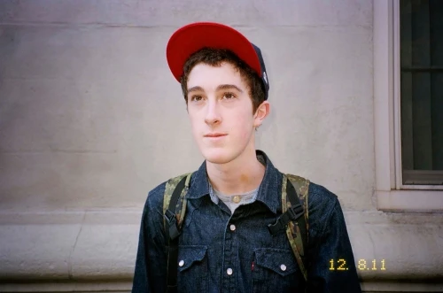 Portrait of a young adult man wearing a cap and backpack, standing in front of a building with a window. Captured in natural light.