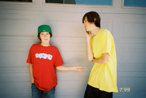 Two teenage boys stand outdoors in front of a gray garage door. One wears a red t-shirt and a green baseball cap, smiling at the other.