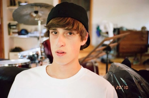 Portrait of a young man in a white t-shirt and black baseball cap, posing indoors with drums and household items in the background.