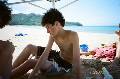 Teenage boy with dark hair sits on a sandy beach near the ocean with other people, a blue bucket, and a red towel.