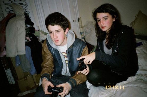 A young man holds a game controller while a young woman points during a video game session on a bed in a casual indoor setting.