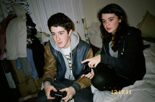 A young man holds a game controller while a young woman points during a video game session on a bed in a casual indoor setting.