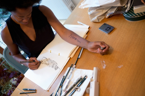 Black woman sketching in a notebook at a table with pens, an MP3 player, and the word 'Heroism' tattooed on her forearm in natural light.