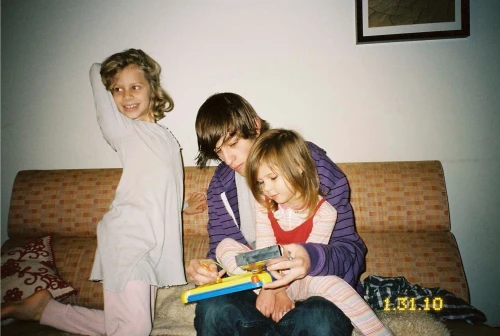 Young man holding a toy while a girl sits on his lap and another girl poses standing on a patterned sofa.