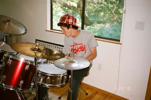 Young adult male playing drums indoors, wearing a hat and grey Stussy tee shirt in a room with a window and natural lighting.