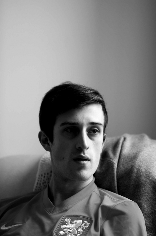 Black and white portrait of a young adult male wearing a sports shirt with a lion emblem, seated indoors with soft lighting.