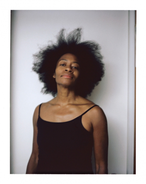 Portrait of a Black woman with voluminous afro hair wearing a black camisole against a plain white wall