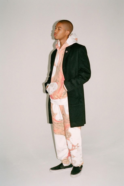 A young adult Black man in a patterned hoodie and pants stands in a photo studio, wearing a dark overcoat and black sneakers in a medium shot.