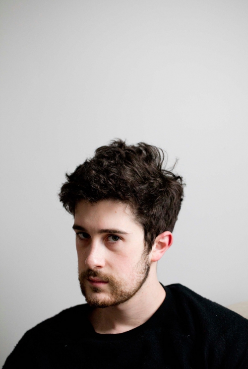 Indoor portrait of a young adult male with dark curly hair and a light beard, wearing a black sweater, against a plain white wall.