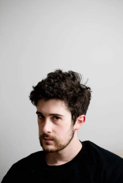 Indoor portrait of a young adult male with dark curly hair and a light beard, wearing a black sweater, against a plain white wall.