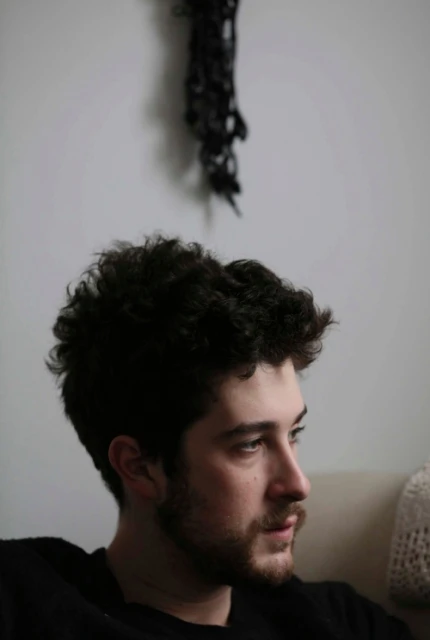 Portrait of a young adult man with dark curly hair and a beard looking off to the side indoors with a neutral facial expression.
