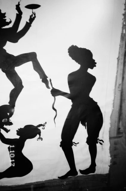 Silhouette of people in an indoor theatrical setting, possibly a shadow play, presenting three figures, one with a snake.