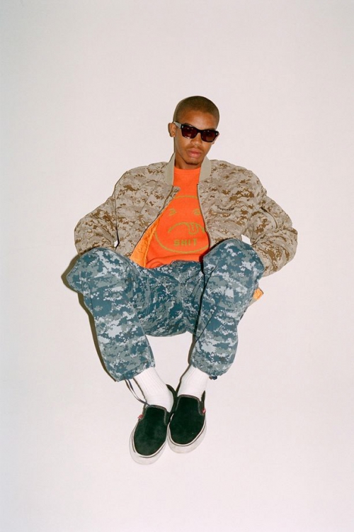 Male fashion model wearing camouflage jacket and pants with black slip-on shoes against a white backdrop in a studio setting.