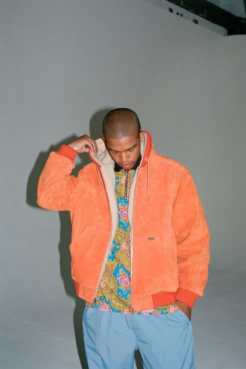 Young man in a stylish orange hooded jacket with blue pants posing in a studio with soft lighting, showcasing contemporary fashion.
