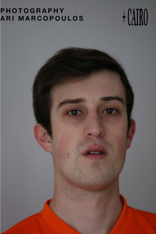 Portrait of a man wearing an orange shirt against a neutral grey background photographed by Ari Marcopoulos.