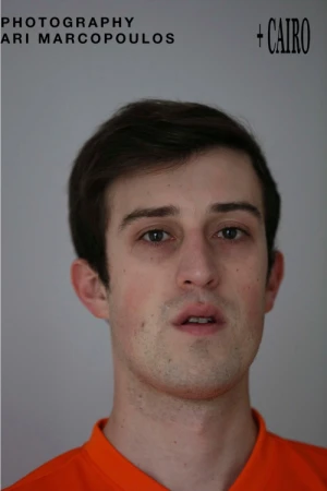 Portrait of a man wearing an orange shirt against a neutral grey background photographed by Ari Marcopoulos.