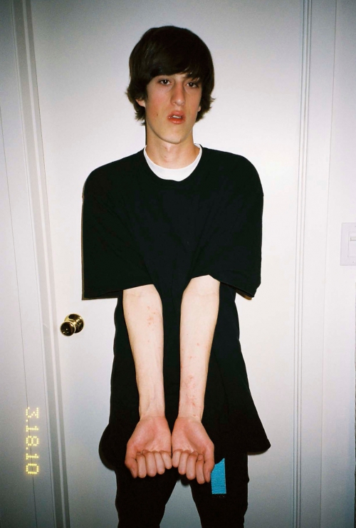 A young adult with brown hair stands indoors wearing a black shirt and pants, facing the camera against a white door backdrop.