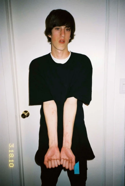 A young adult with brown hair stands indoors wearing a black shirt and pants, facing the camera against a white door backdrop.