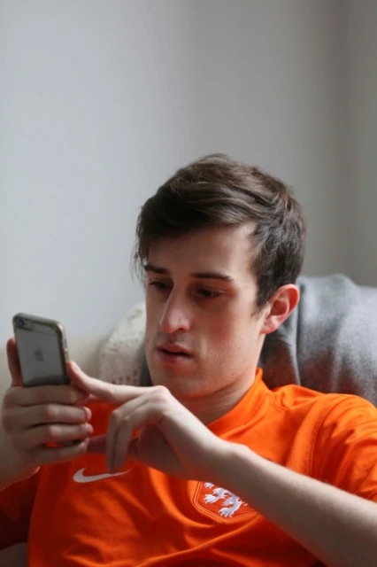 Young man in an orange shirt holds a cell phone indoors, looking at the screen with a focused expression. He's seated and relaxed.