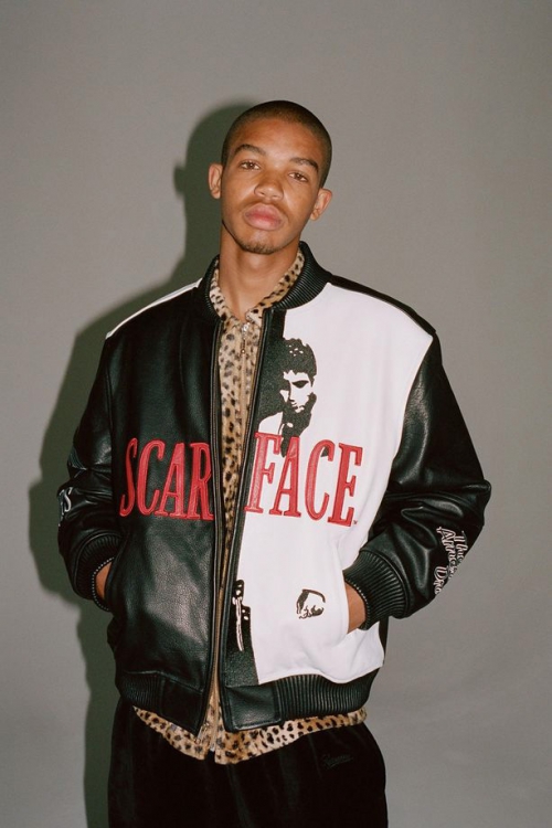 Portrait of a young adult man wearing a Scarface-themed leather bomber jacket and animal print shirt in a studio setting.