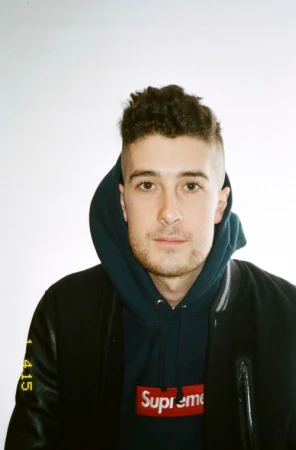 Portrait of a young man wearing a dark hoodie with a red logo and a dark bomber jacket against a plain white background.