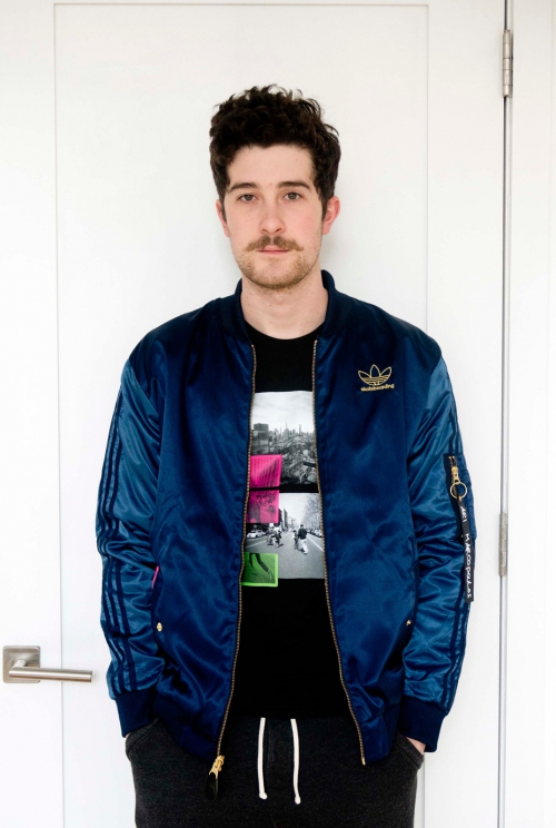 Portrait of a dark-haired man in a blue satin bomber jacket, black tee shirt with graphic, and grey sweatpants standing against a white door.