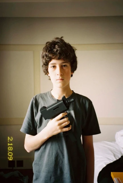 Boy holding a toy gun in an indoor environment, wearing a grey t-shirt, and standing in front of a white wall. Focus is on the subject.