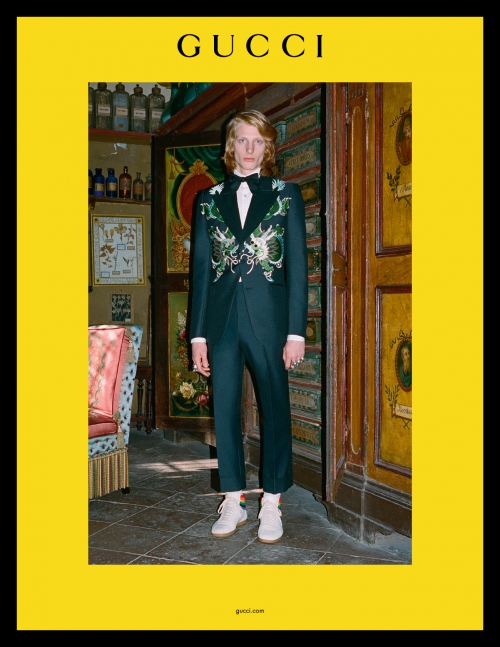 Male fashion model wears an embroidered Gucci suit and white sneakers while posing indoors against an ornate door and cabinet.