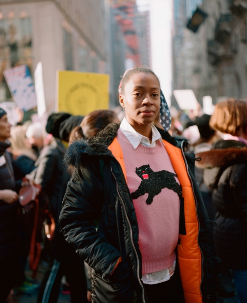 A young woman in a pink sweater with a black cat design stands on a city street amongst a crowd, wearing a black jacket with orange lining.