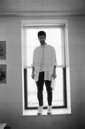 Monochrome portrait of a man standing in a window holding a notebook, photographed by Ari Marcopoulos in New York.