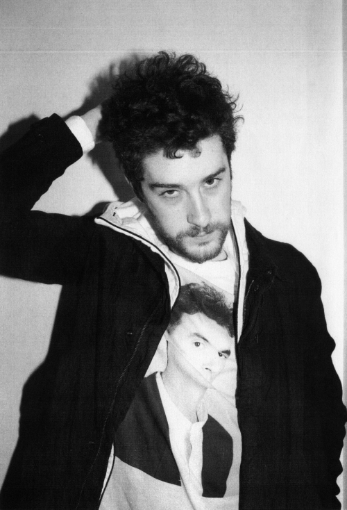 Monochrome portrait of an adult male with curly hair wearing a jacket over a t-shirt with a printed face, indoor setting, white background.