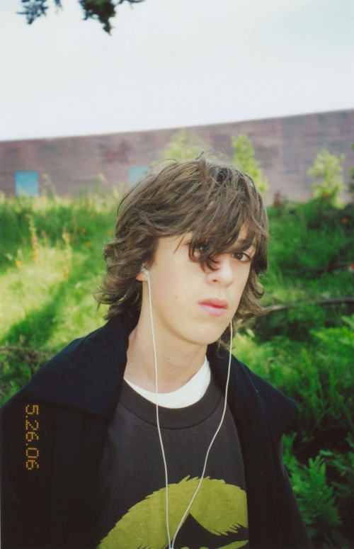 Portrait of a young adult wearing headphones, a graphic t-shirt, and a dark coat outdoors, standing in front of green foliage.