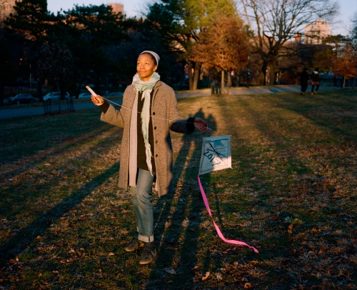 A woman in a grey coat and jeans stands in a grassy park holding a kite on a sunny day. Trees and buildings are in the background.