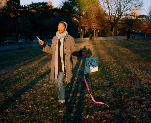 A woman in a grey coat and jeans stands in a grassy park holding a kite on a sunny day. Trees and buildings are in the background.