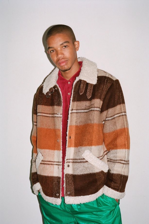 Man in a brown and tan plaid jacket with a white faux fur collar and green pants poses against a white backdrop in a studio shot.