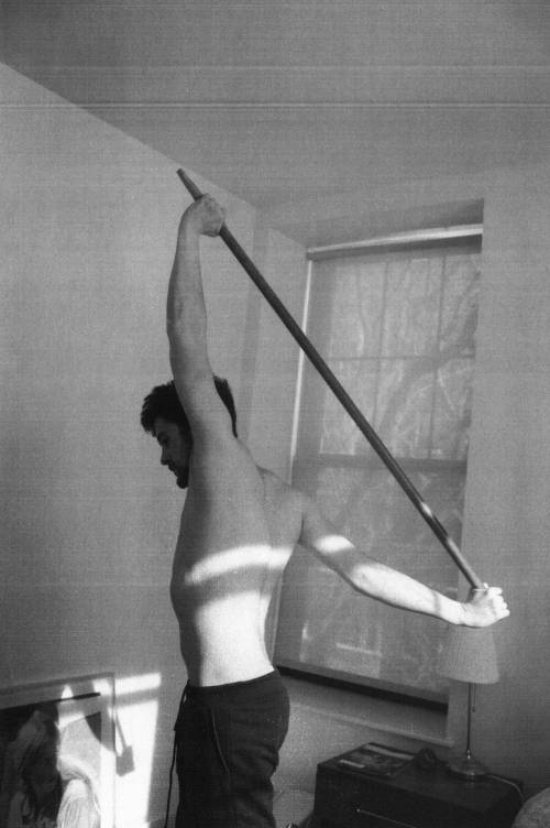 Shirtless man doing shoulder stretch with wooden pole in indoor setting with natural window light in black and white image.