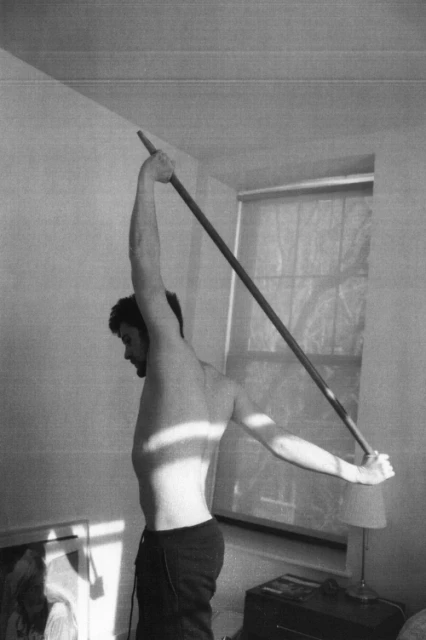 Shirtless man doing shoulder stretch with wooden pole in indoor setting with natural window light in black and white image.