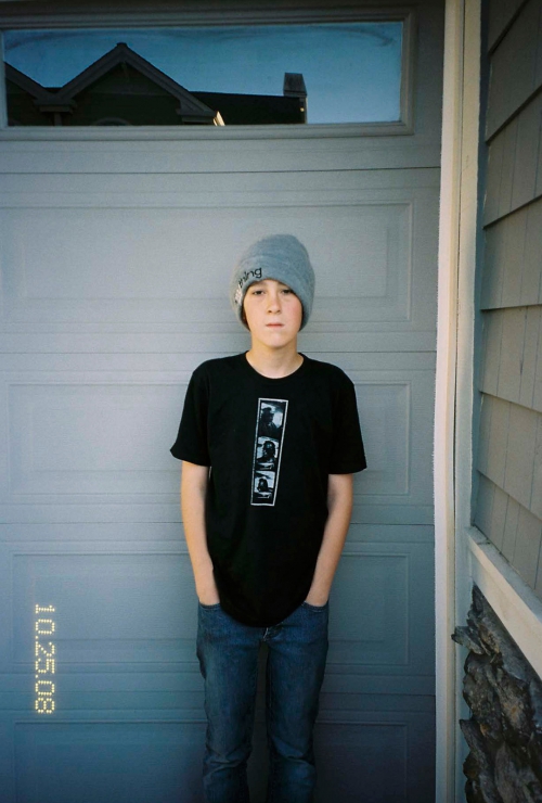 Teenager stands outdoors in front of a gray garage door wearing a beanie, black graphic tee, and blue jeans with his hands in his pockets.