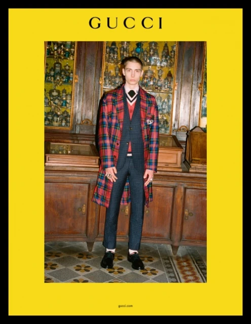 A young male model poses indoors in a Gucci ad, wearing a patterned suit, plaid coat, and tie, against a backdrop of display cases.