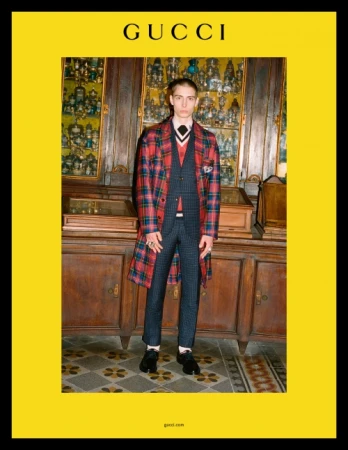 A young male model poses indoors in a Gucci ad, wearing a patterned suit, plaid coat, and tie, against a backdrop of display cases.