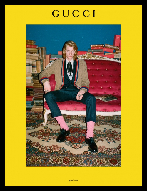 Fashion shot: Young adult man poses seated on a red sofa in a suit with a cardigan, with a backdrop of books and Gucci logo above.