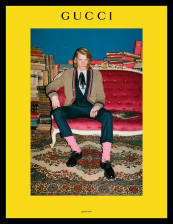 Fashion shot: Young adult man poses seated on a red sofa in a suit with a cardigan, with a backdrop of books and Gucci logo above.