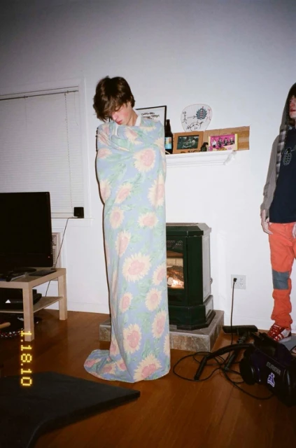 A person wrapped in a floral blanket standing in a living room, with a second person visible in the background.