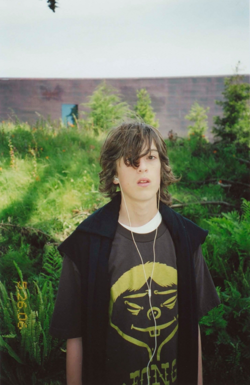 Young person with long dark hair wearing a graphic t-shirt and white earphones standing outdoors in lush greenery.